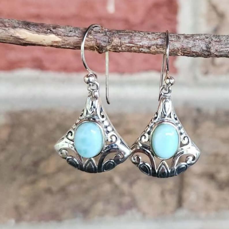 May include: A pair of silver earrings with a light blue oval stone set in a decorative, openwork design. The earrings are suspended from a curved hook and are displayed against a blurred background.