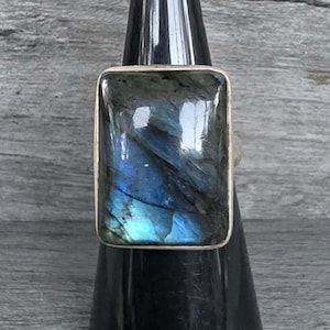 May include: A rectangular labradorite gemstone ring featuring a polished, smooth surface with iridescent blue and grey flashes set within a simple metal frame. The stone displays natural internal fractures and deep color variations across its face.