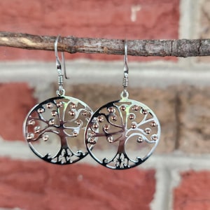 May include: A pair of silver-colored dangle earrings. Each earring features a circular design with a tree of life motif. The tree's branches and leaves are accented with small, copper-colored beads. The earrings hang from silver hooks.
