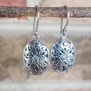 May include: A pair of silver drop earrings featuring an oval-shaped design with intricate, openwork filigree patterns. The metal has a polished finish with darkened recessed areas to highlight the swirling, floral-inspired scrollwork details.