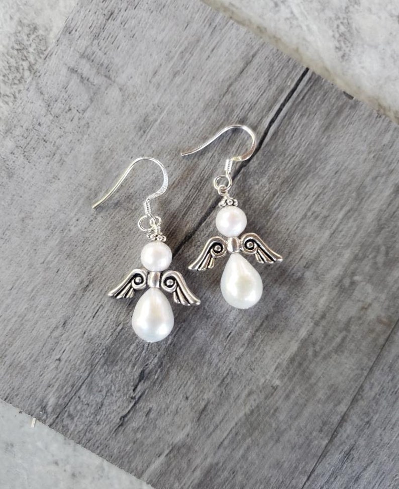 Puede incluir: Un par de pendientes de plata con forma de &aacute;ngel con detalles de perlas blancas. Los &aacute;ngeles tienen alas delicadas y las perlas son de forma de l&aacute;grima.