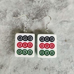 May include: A pair of white square earrings with a Mahjong tile design. Each earring features three rows of three circles in black, red, and green. The earrings have silver-colored fish hook closures.