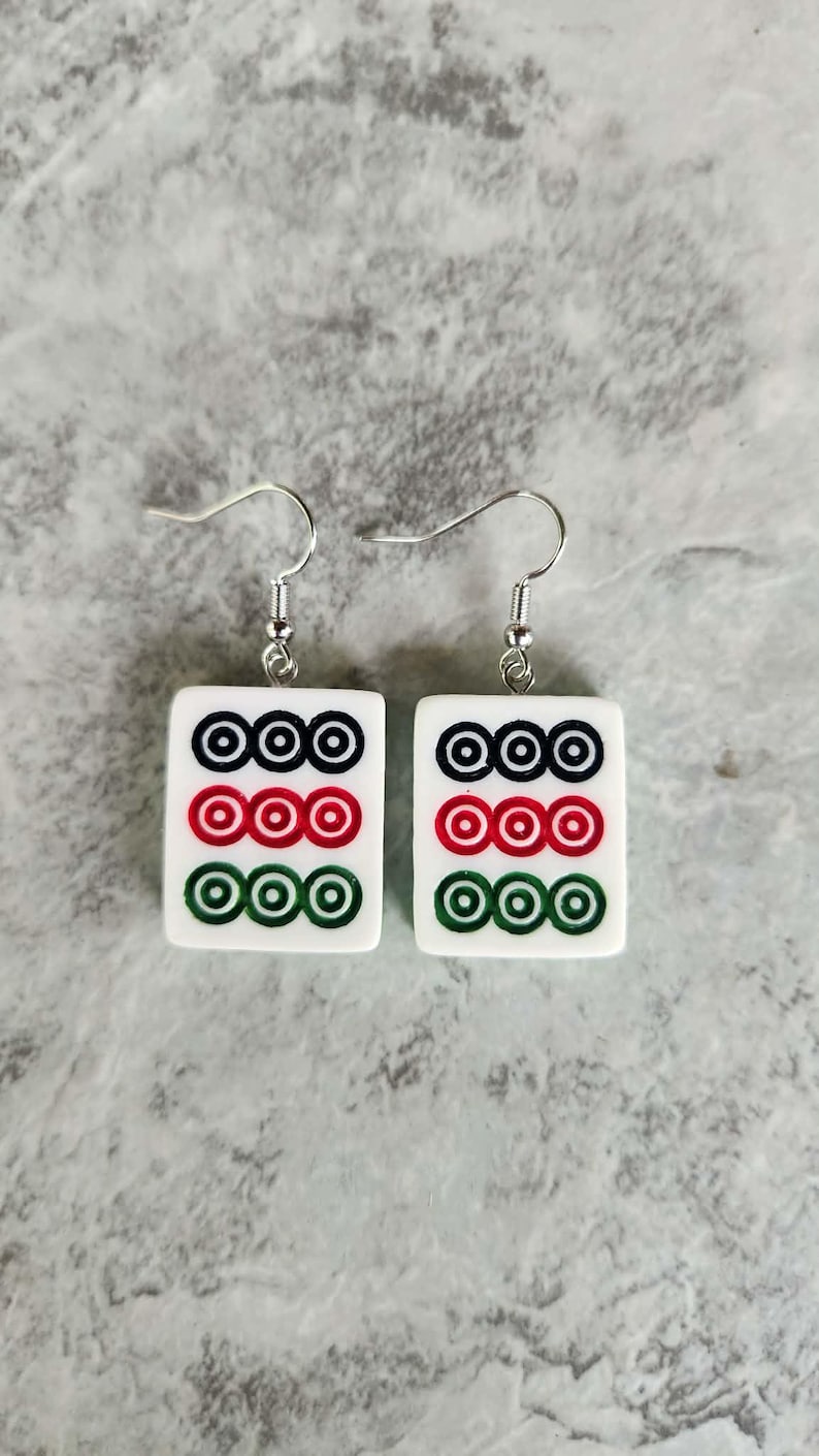 May include: A pair of white square earrings with a Mahjong tile design. Each earring features three rows of three circles in black, red, and green. The earrings have silver-colored fish hook closures.