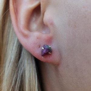 May include: A pair of small, raw ruby earrings with gold settings. The earrings are worn in the earlobe of a person with blonde hair.