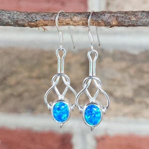 May include: A pair of silver drop earrings featuring oval-shaped blue opal cabochons set within a decorative, open-work metal frame. The earrings hang from simple silver hooks, showcasing a polished finish and vibrant, iridescent blue tones.