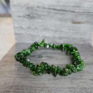 May include: A green beaded bracelet made of small, irregularly shaped stones. The bracelet is a vibrant shade of green and rests on a wooden surface. The stones appear to be polished and have a slight sheen.