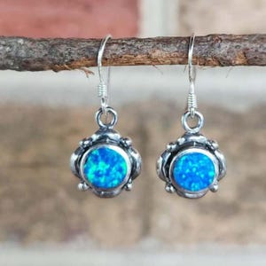 May include: A pair of silver drop earrings featuring round, vibrant blue opal cabochons set within a decorative, scalloped metal frame. Each earring hangs from a simple silver hook, showcasing a bright, iridescent blue stone with a textured finish.