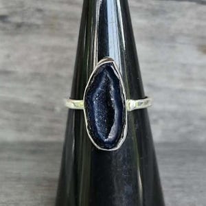 May include: A silver-toned ring featuring an elongated, irregular-shaped dark blue druzy gemstone centerpiece. The stone has a textured, crystalline surface with a deep navy hue, set within a smooth metallic band that encircles the finger.