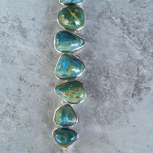 May include: A silver bracelet featuring seven polished, irregular, cabochon-cut stones in shades of blue, green, and brown. The stones are set in silver bezels and connected by silver links, finished with a toggle clasp and a short chain.