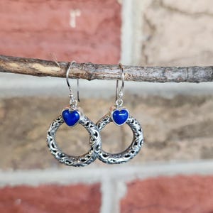 Puede incluir: Pendientes de plata con una piedra azul en forma de coraz&oacute;n y un dise&ntilde;o de filigrana circular. Los pendientes cuelgan de una ramita, con un fondo de pared de ladrillo.