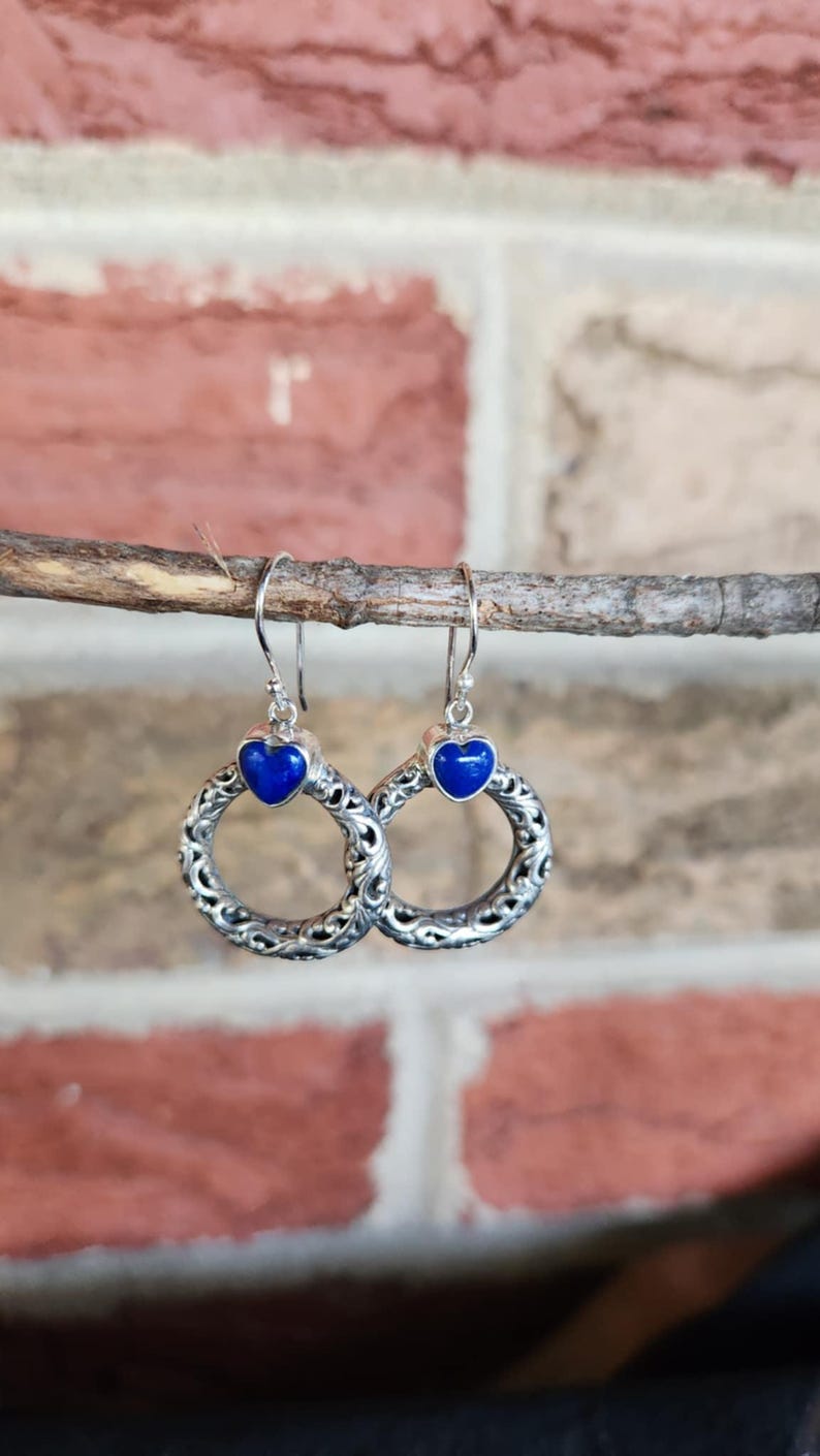 Puede incluir: Pendientes de plata con una piedra azul en forma de coraz&oacute;n y un dise&ntilde;o de filigrana circular. Los pendientes cuelgan de una ramita, con un fondo de pared de ladrillo.