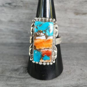 May include: A rectangular sterling silver ring featuring a multi-colored inlay of turquoise, white, and orange stone segments. The stone setting is framed by a beaded silver border and mounted on a polished silver band with decorative side details.