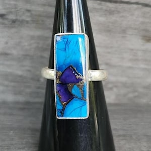 May include: A rectangular ring featuring a vibrant blue and deep purple stone inlay with dark veining. The stone is set in a polished silver-toned metal band. The design showcases a mosaic-like pattern of contrasting colors held within a smooth, metallic frame that encircles the finger.