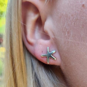 May include: A close-up of a person's ear with a silver starfish stud earring. The earring is a simple, five-pointed starfish design. The person has blonde hair.