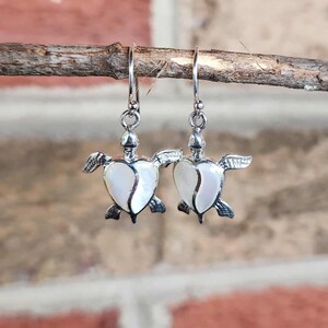 May include: A pair of silver-toned turtle earrings. Each earring features a turtle design with a white, heart-shaped shell. The earrings are suspended from a silver hook and are displayed against a brick background.
