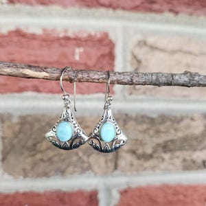 May include: A pair of silver-toned earrings with a triangular design, featuring a light blue, oval-shaped stone in the center. The earrings are suspended from a thin wire and are displayed against a brick background.
