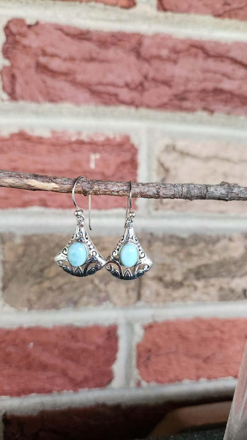May include: A pair of silver-toned earrings with a triangular design, featuring a light blue, oval-shaped stone in the center. The earrings are suspended from a thin wire and are displayed against a brick background.