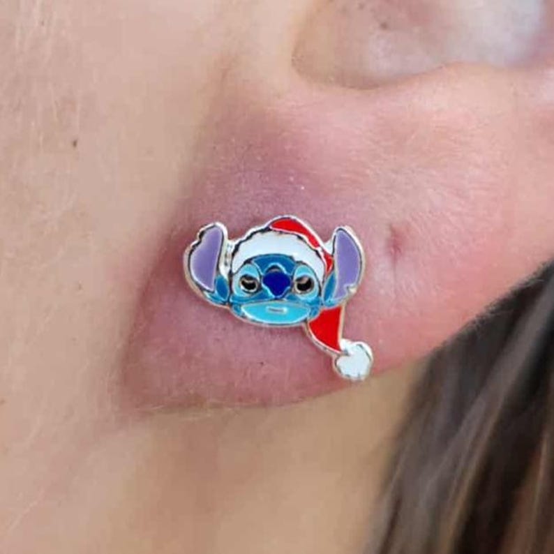 May include: A silver-toned stud earring featuring a blue cartoon character wearing a red and white Santa hat. The character has large ears and a light blue face. The earring is a festive accessory.
