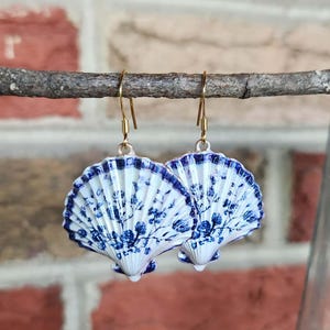 May include: A pair of dangle earrings featuring white scallop shells decorated with a blue floral pattern. The shells are suspended from gold-toned metal hooks. The design on the shells mimics a classic porcelain aesthetic with intricate blue botanical illustrations against a white background.