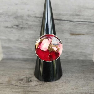 May include: A round ring with a silver band and a large, colorful stone. The stone features a mix of red, pink, gold, and white fragments, creating a mosaic-like appearance. The ring is displayed on a black cone-shaped stand.