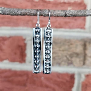 May include: A pair of silver dangle earrings. Each earring features a rectangular design with a row of silver beads down the center. The earrings are suspended from a silver hook. The background is a blurred brick wall.
