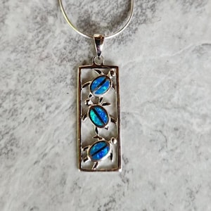 May include: Silver pendant necklace featuring three turtle designs with blue opal-like inlays. The turtles are set within a rectangular frame, suspended from a silver chain. The pendant is displayed against a textured, light gray surface.