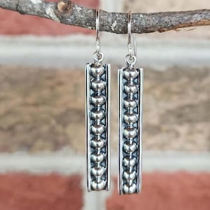 May include: A pair of silver dangle earrings featuring a long, rectangular frame. Each earring contains a vertical row of small, polished silver spheres set within the center of the frame, suspended from a simple silver hook wire attachment.