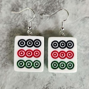 May include: A pair of white earrings with a silver hook. Each earring features a rectangular tile with three rows of concentric circles in black, red, and green. The earrings are designed to resemble Mahjong tiles.