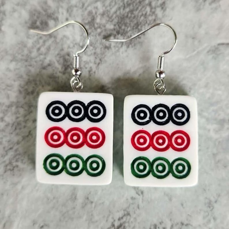 May include: A pair of white earrings with a silver hook. Each earring features a rectangular tile with three rows of concentric circles in black, red, and green. The earrings are designed to resemble Mahjong tiles.
