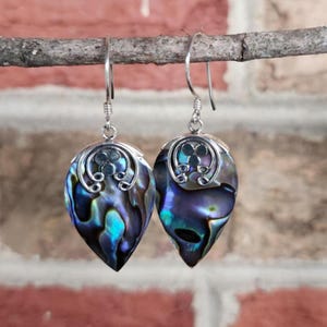 May include: A pair of teardrop-shaped earrings with iridescent abalone shells. The earrings have silver-colored metalwork with decorative swirls and hooks. The abalone shells display a mix of blue, green, and purple hues, creating a shimmering effect.