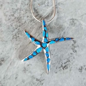 May include: A silver starfish pendant with a bright blue, opal-like inlay. The five-pointed starfish is suspended from a thin silver chain. The pendant is set against a mottled gray background.