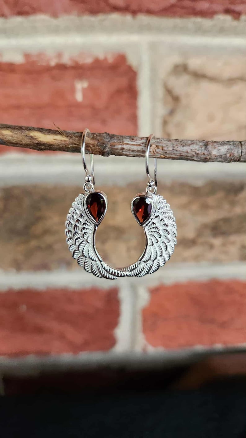 May include: Silver dangle earrings with a wing design. Each earring features a teardrop-shaped, deep red gemstone at the top and a detailed wing shape below. The earrings are displayed against a brick background.