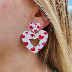 May include: A pair of white heart-shaped earrings with a red heart pattern. The earrings are layered, with a smaller heart on top of a larger heart.