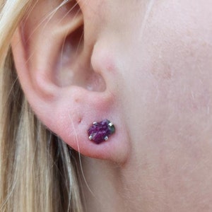May include: A close-up of a person's ear with a small, raw ruby stud earring. The earring is a deep pinkish-red color, set in a silver bezel. The person has blonde hair.