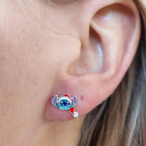 May include: A pair of small, colorful stud earrings. Each earring features a blue alien character wearing a red Santa hat. The earrings are set against the skin of a person's ear.