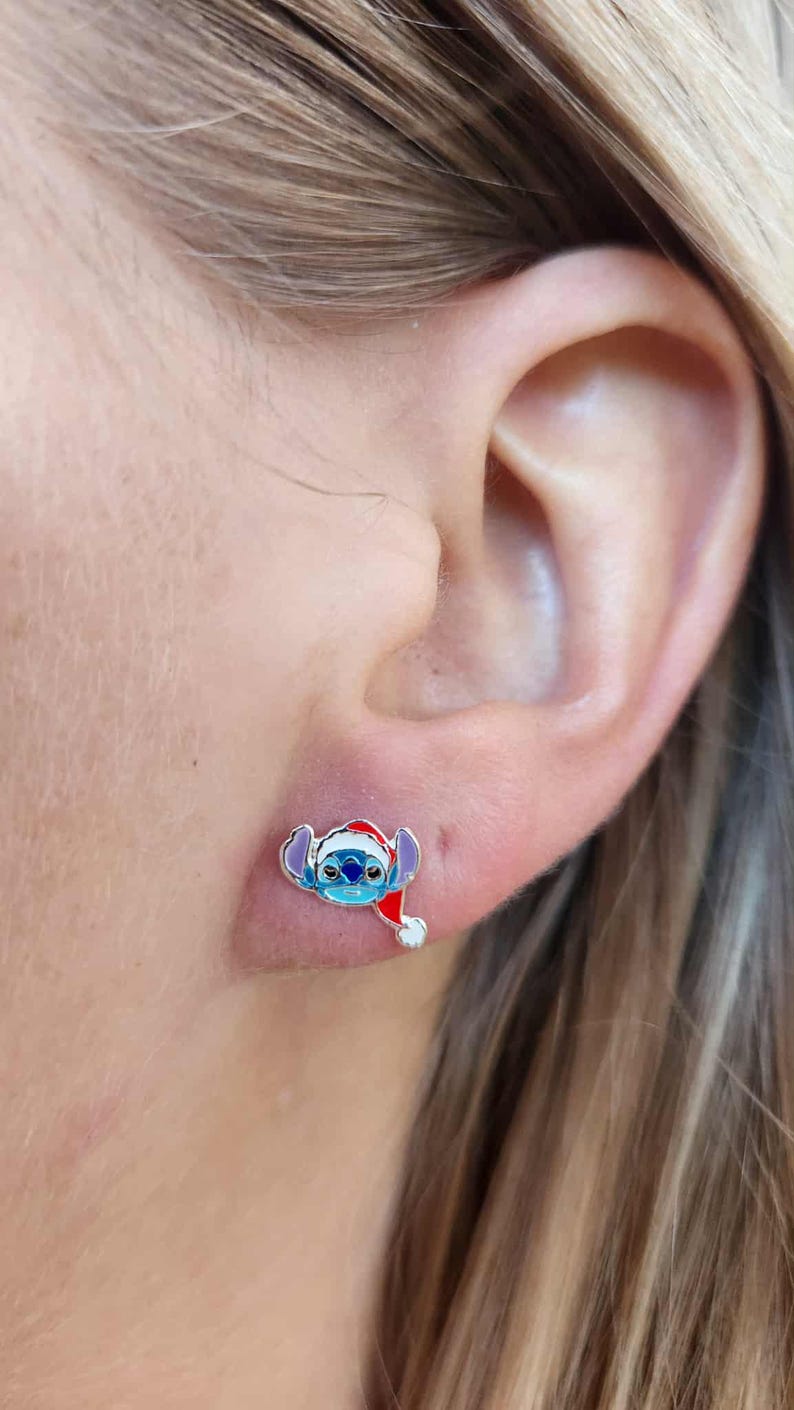 May include: A pair of small, colorful stud earrings. Each earring features a blue alien character wearing a red Santa hat. The earrings are set against the skin of a person's ear.