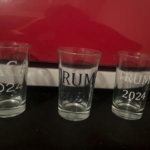 May include: Three clear glass shot glasses with white lettering. The first glass says "MAGA 2024", the second glass says "TRUMP 2024", and the third glass says "TRUMP 2024".