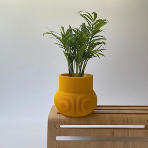 Pode incluir: Um vaso amarelo vibrante com uma textura vertical canelada distinta e uma base arredondada, contendo uma palmeira-leque verde. O vaso apresenta uma secção superior cilíndrica larga que se estreita para um corpo inferior bulboso e ranhurado.