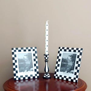May include: A decorative arrangement featuring two black and white chequered picture frames, a black and white candlestick holder, and a white candle with black polka dots. The frames display black and white landscape photos.