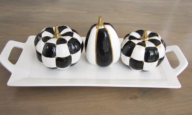 Op de afbeelding: Drie decoratieve pompoenen met zwart-witte patronen en gouden stelen, gerangschikt op een wit rechthoekig dienblad met handvatten. Twee pompoenen hebben een ruitpatroon, terwijl de middelste pompoen verticale zwart-witte strepen heeft.