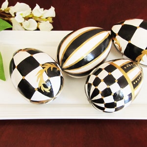 Peut inclure: Cinq œufs de Pâques décoratifs sur un plateau rectangulaire blanc. Les œufs sont peints en noir et blanc avec des accents dorés. Les œufs ont des motifs différents, notamment des rayures, des damiers et des motifs géométriques.