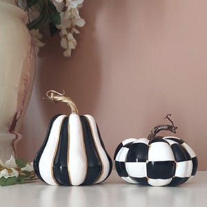 May include: Two decorative pumpkins featuring black and white patterns. The taller pumpkin has vertical stripes, while the shorter, rounder pumpkin has a checkered pattern. Both pumpkins are accented with gold paint and have metallic gold stems.