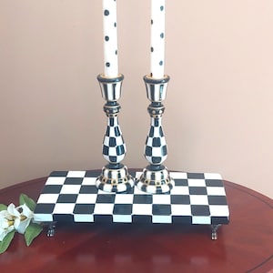 May include: Two tall candlesticks with a black and white checkered pattern and gold accents, each holding a white candle with black polka dots. They stand on a rectangular black and white checkered riser with four small silver-toned feet.