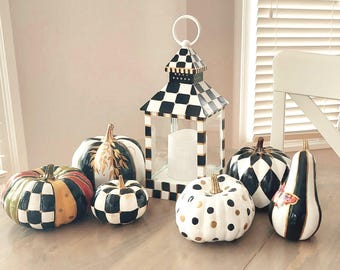 May include: A decorative lantern with a black and white checkered pattern and a collection of six pumpkins. The pumpkins feature various designs including black and white checks, gold polka dots, solid black, and painted gold leaf accents.