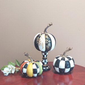 May include: Three decorative pumpkins featuring black and white chequered patterns. One pumpkin sits on a pedestal base, another is round with black and white checks, and the third features yellow, orange, and green segments with black and white checks.
