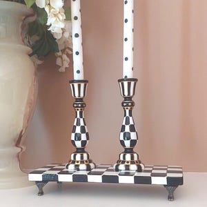 May include: Two tall candlesticks with black and white checkered patterns and gold accents, each holding a white taper candle with black polka dots. The candlesticks stand on a rectangular black and white checkered base with four small metal feet.