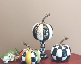 Peut inclure: Trois citrouilles décoratives en céramique. L'une est à carreaux noirs et blancs, une autre est rayée avec des accents de feuilles d'or, et la troisième est multicolore. Toutes ont des tiges incurvées et sont posées sur une table rouge.