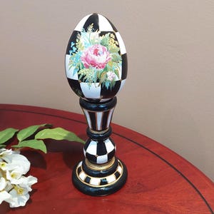 Peut inclure: Objet décoratif en forme d'œuf avec un motif à carreaux noir et blanc. L'œuf présente un motif floral avec des fleurs roses et bleues. Il est monté sur un piédestal noir et or, créant un contraste saisissant.