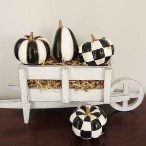 Op de afbeelding: Een witte houten kruiwagen gevuld met beige papieropvulling en drie decoratieve pompoenen met zwart-witte strepen en een ruitpatroon met gouden stelen. Een vierde bijpassende pompoen met ruitpatroon staat voor de kruiwagen.