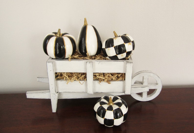 Op de afbeelding: Een witte houten kruiwagen gevuld met beige papieropvulling en drie decoratieve pompoenen met zwart-witte strepen en een ruitpatroon met gouden stelen. Een vierde bijpassende pompoen met ruitpatroon staat voor de kruiwagen.